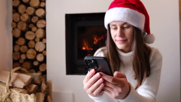 有选择地关注微笑的年轻女性在家里用手机发短信庆祝圣诞节 千禧年女性寒假期间在壁炉附近有智能手机呼叫 传播概念 — 图库视频影像