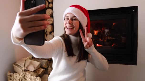 快乐的小女孩戴着圣诞礼帽摆出完美的笑容 坐在壁炉边温暖的地板上 用智能手机拍照 在电视上聊天 新年远程庆祝活动 — 图库视频影像
