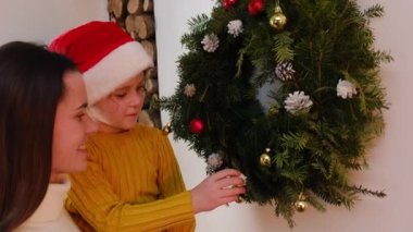 Kırmızı şapkalı, şefkatli, genç ve güzel bir kızın Noel çelengini süsleyip evde şöminenin yanında duran güzel kızına yaklaş. Noel ve yeni yıl tatili konsepti