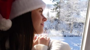 Beyaz örgü süveter ve Noel Baba kırmızı şapkalı mutlu, rüya gören genç bir kadın pencere kenarında oturmuş güzel, karlı, karışık ormanlara bakıyor. Şirin bayan tatil haftasonunun tadını çıkarıyor.