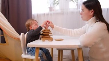 Mutlu genç anne ve tatlı bebek kız tahta bloklarla gülerek çocuk odasındaki beyaz masada oturuyor, neşeli anne küçük kızla eğleniyor evde eğlenceli aktivitelerin tadını çıkarıyor birlikte gülüyorlar.