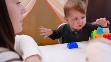 Genç anne ve tatlı bebek, rahat oturma odasındaki masada plastik küplerle oynuyorlar. Güzel bir aile oyun zamanı inşaatında renkli tuğlalar kullanmaktan zevk alır. Hafta sonu eğlence etkinliği konsepti