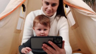 Modern teknoloji konseptine bağımlı. Mutlu bir anne ve güzel bir kız çocuğu cep telefonuyla eğleniyor. Genç dadı, sevimli küçük kızıyla mobil oyunlar oynuyor. Evdeki rahat çadırda oturuyor.