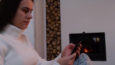 Evdeki rahat koltukta oturan, şöminenin yanındaki yazlık evde dinlenen, akıllı telefon cihazıyla vakit geçiren, internette sohbet eden, internetten alışveriş yapan güzel bir genç kadının portresi.