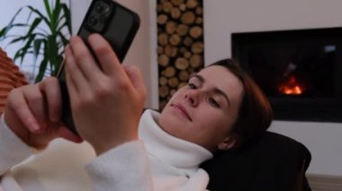 Modern köy evindeki rahat bir kanepede uzanan güzel ve çekici bir kadının yan görüntüsü. Şömineli rahat. Telefon uygulamaları kullanarak akıllı telefonu tut.