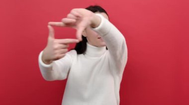 Genç esmer kadının portresi kameraya el çerçevesinden bakar, fotoğraf çeker ya da hedefe odaklanır, beyaz örgü süveter giyer, kırmızı renkli arka plan stüdyosunda izole bir şekilde poz verir.