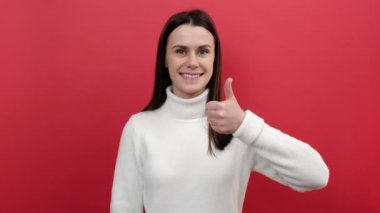 Mutlu beyaz kadın kameraya bakıyor dişlek gülüşü ve baş parmaklarını gösteriyor, onay işareti, iyi geri bildirim, rahat örgü süveter giyiyor, stüdyoda pastel kırmızısı arka planda izole edilmiş.