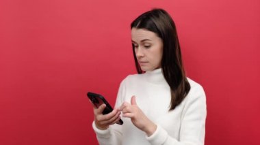 Cep telefonu kullanan güzel, esmer bir kadının portresi, kayan ağ gibi gülümseyen, mobil uygulamadan tatmin olmuş, rahat beyaz bir süveter giyen, stüdyoda kırmızı arka planda izole bir şekilde poz veren