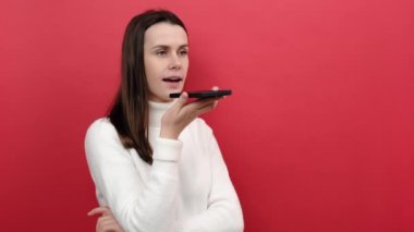İyimser genç esmer kadının portresi cep telefonuyla ses kaydı yapıyor, akıllı cihazla konuşuyor, rahat beyaz örgü süveter giyiyor, kırmızı renkli arka plan duvarında izole edilmiş poz veriyor.