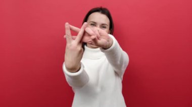 Güzel ve güzel bir kadın hashtag işareti yapmak için parmaklarını çapraz yapıyor ve kameraya dişlek gülüşüyle bakıyor, beyaz örgü süveter giyiyor, kırmızı arka planda izole bir şekilde poz veriyor.
