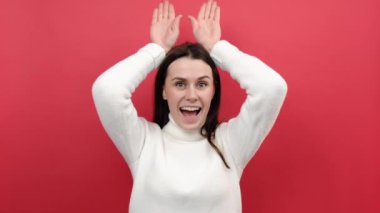 Neşeli, neşeli, genç bir kadın başının üstünde el ele tutuşuyor. Tavşan kulakları, flört, paskalya sembolü, beyaz örgü süveter giymiş, kırmızı arka plan stüdyosuna izole edilmiş bir şekilde poz veriyor.