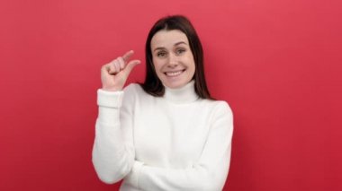 Genç esmer kadının portresi parmaklarıyla küçük boy gösteriyor, santimetre boyunda, beyaz süveter giyiyor, kırmızı stüdyo arka planında izole edilmiş poz veriyor. Ölçek kavramı ölçülüyor
