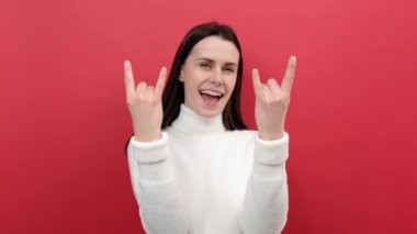 Mutlu genç kadın portresi çılgın hissediyor, şeytan boynuzlarını, rock and roll el hareketlerini gösteriyor, başarıya seviniyor, beyaz kazak giyiyor, stüdyonun kırmızı arka plan duvarında izole edilmiş poz veriyor.