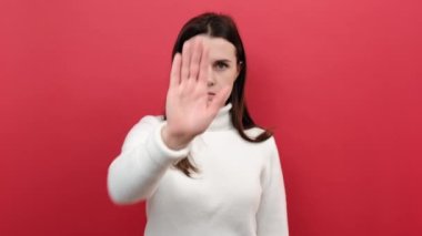 Duygusal genç bir kadın, hayır diyerek, hayır diyerek, beyaz süveter giymiş, kırmızı arka planda izole edilmiş, inkar ya da kısıtlamayı ifade ederek jesti durduruyor. Olumsuz insani duygular, duygular
