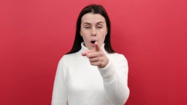 Kamerayı işaret eden genç esmer kadın portresi, şok olmuş büyük gözlerle bakıyor, seçim yapıyor ve şaşırıyor, örgü süveter giymiş, kırmızı arka plan stüdyosunun üzerine izole edilmiş poz veriyor.