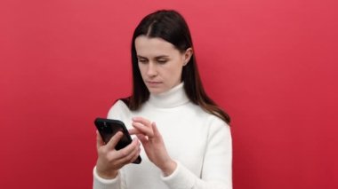 Şok edici haberleri duyduktan sonra şaşırmış bir şekilde cep telefonuyla konuşan çekici genç bir kadının portresi, stüdyoda kırmızı arka plan duvarının üzerine izole edilmiş beyaz bir süveter giyiyor.