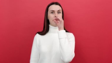 Tatlı milenyum bayanının portresi ağzını zip jest ile kapatıyor, sonsuza kadar sır saklayacağına söz veriyor, beyaz örgü süveter giyiyor, stüdyoda pastel kırmızısı arka plan duvarının üzerine izole edilmiş poz veriyor.