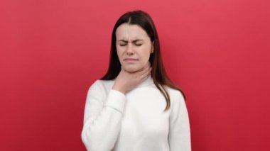 Acı içinde kıvranan, boğaz ağrısı çeken, boğaz ağrısı çeken, virüs ya da bademcik iltihabı olan 20 'li yaşlardaki mutsuz genç kadın portresi beyaz süveter giyer, stüdyoda kırmızı arka plan duvarının üzerine izole bir şekilde poz verir.