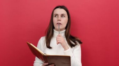 Düşünceli, genç, esmer bir kadının portresi notlar alıyor, kalemle not defterine düşünceler yazıyor, liste yapıyor, beyaz örgü süveter giyiyor, pastel kırmızısı arka plan stüdyosuna izole edilmiş poz veriyor.