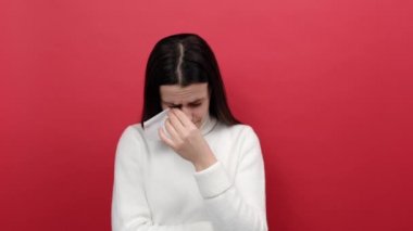 Üzgün genç esmer kadın portresi dramatik bir şekilde ağlıyor, üzüntü duyuyor, umutsuz duygular içinde, beyaz örülmüş süveter giymiş, kırmızı arka plan duvarında izole bir şekilde poz veriyor.