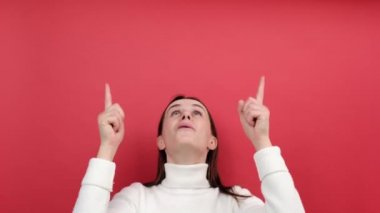 Heyecanlı gülümseyen genç bir kadının portresi reklam içeriği için boş bir yere işaret ediyor ve şaşırdığını ifade ediyor, beyaz örgü süveter giyiyor, stüdyoda kırmızı renkli arka planda izole bir şekilde poz veriyor.