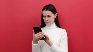 Genç esmer kadın portresi cep telefonu kullanarak sosyal medyaya kayıyor, cep telefonu uygulamasına şaşırıyor, örgü süveter giyiyor, kırmızı arka plan stüdyosunda izole bir şekilde poz veriyor.