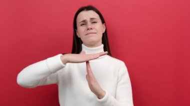 Ciddi genç bir kadının portresi el hareketi yapıyor, yalvararak bakıyor, teslim tarihinden endişe ediyor, beyaz örgü süveter giyiyor, stüdyoda kırmızı arka plan duvarına izole edilmiş poz veriyor.