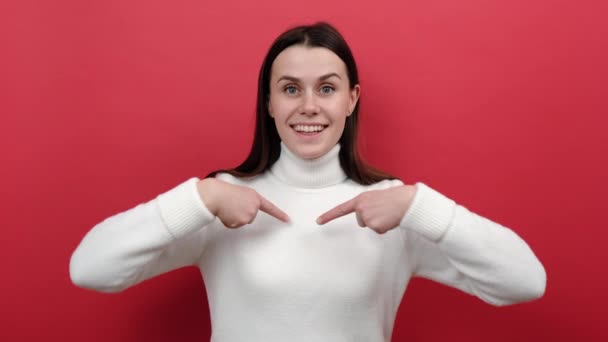 Excited young female pointing herself and looking at camera with shocked expression, astonished by sudden success, dressed in white knitted sweater, posing isolated over red color background studio