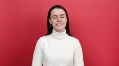 20 'li yaşlarda, gözleri kapalı, yastık ve uyku gibi kolları başında duran, rahat, beyaz örülü süveter giyen, kırmızı arka plan stüdyosunun üzerine izole bir şekilde poz veren, tembel bir genç kadın portresi.