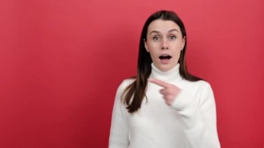 20 'li yaşlarda beyaz bir kadın reklamınız için boş bir yeri işaret ediyor. Ulaşılabilirliğini merak ediyor, beyaz örgü süveter giyiyor, kırmızı renkli arka plan stüdyosuna izole edilmiş poz veriyor.