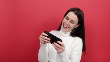 Cep telefonuyla video oyunu oynayan mutlu beyaz kadın portresi, gülümseyen ve oyun oynamaktan duygusal olarak keyif alan, örgü ören süveter giyen, stüdyoda kırmızı arka plan duvarının üzerinde izole bir şekilde poz veren