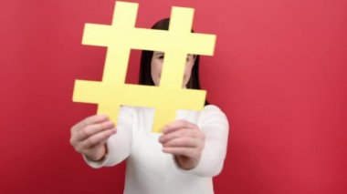Mutlu sevimli genç kadın sarı hashtag sembolünü işaret ediyor, foruma katılmayı öneriyor, kırmızı stüdyo arka planında izole edilmiş rahat beyaz bir kazak giyiyor. Popüler blog fikri kavramı