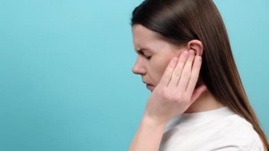 Kulak ağrısı ve kulak çınlamasından muzdarip üzgün genç kadını kapatın, mavi arka planda izole edilmiş. Kulak ağrısının nedeni kulak iltihabı, kulak kiri, yabancı cisim, sinüs enfeksiyonu veya hava basıncı değişimi.