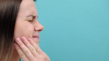 Mavi stüdyo arka planında izole edilmiş, diş ağrısından muzdarip esmer bir kadının yakın çekimi. Diş ağrısının nedenleri diş çürüğü, iltihap, diş apsesi, diş eti hastalığı veya hassas dişlerdir.