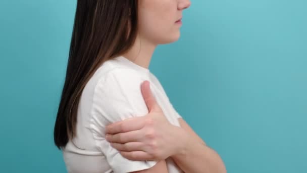 Close up of unhappy sad young female in white t-shirt suffering from arm and shoulder pain, isolated over blue studio background with copy space. Office syndrome. Health care and medical concept