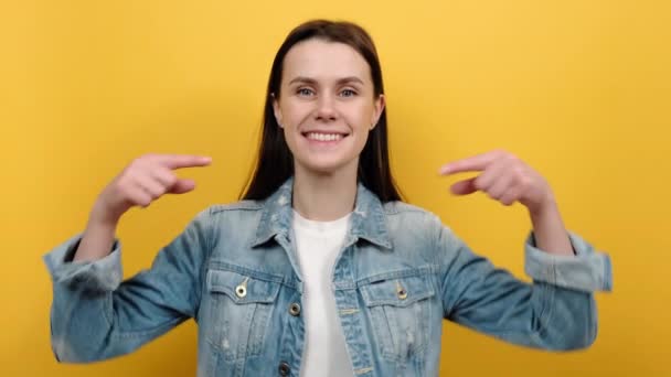 年轻快乐女子的画像 20多岁 手指垂下 要求订阅并喜欢她的帖子 推荐广告区 穿着斜纹棉布夹克 在黄色背景工作室上摆出孤立的姿势 — 图库视频影像