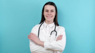 Kendine güvenen, gülümseyen kadın doktor beyaz tıbbi ceket giyer, steteskop mavi stüdyo arka planında duran kameraya bakar. Genç kadın sağlık uzmanı doktor portresi.