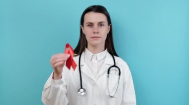 Doktor kadın kırmızı kurdele tutuyor, kameraya bakıyor üniforma ve steteskop giyiyor, mavi arka plan stüdyosunda izole halde. Kan nakli ve bağış. Hemofili, sağlık konsepti. Dünya AIDS ve HIV Günü