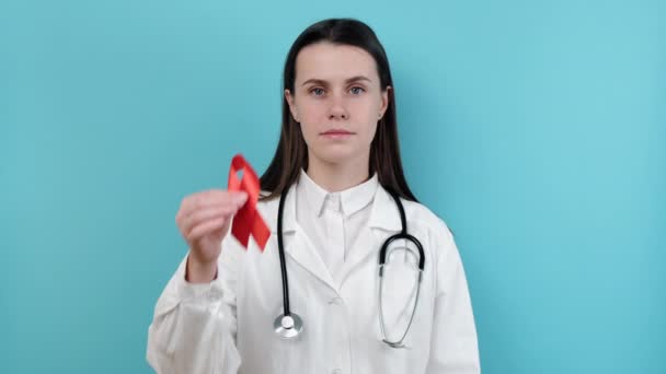 医師の女性は カメラを見て 青い背景スタジオに隔離された均一な聴診器を身に着けている赤いリボンを保持しています 輸血と寄付 血友病 健康概念 世界エイズとHivの日 — ストック動画