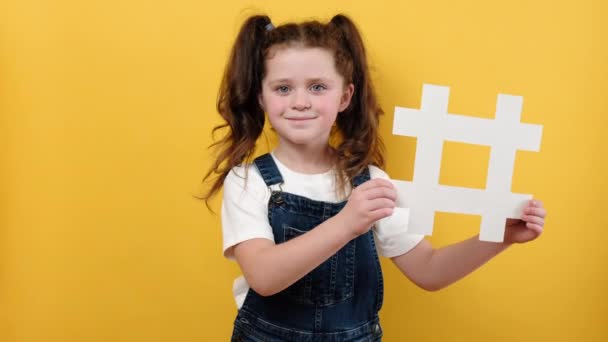 白いハッシュタグのシンボルを持つかわいいかわいい子供の女の子の肖像画 カメラを見て幸せな 黄色の色の背景の壁に隔離されたハッシュ記号を示しています 流行のソーシャルメディアの投稿やブログの概念 — ストック動画