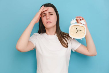20 'li yaşlarda, kafasında çalar saat tutan, kafasına dokunan, beyaz tişört giyen, stüdyoda mavi arkaplan duvarına izole bir poz veren yorgun, üzgün, genç beyaz kadın portresi.