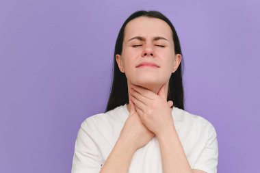 Mor stüdyo duvarında izole edilmiş boğazı ağrıyan genç bir kadına yakın durun. Boğaz ağrısının nedenleri arasında grip, nezle, bakteriyel enfeksiyonlar, zatürree veya alerjiler yer alıyor. Tıbbi konsept