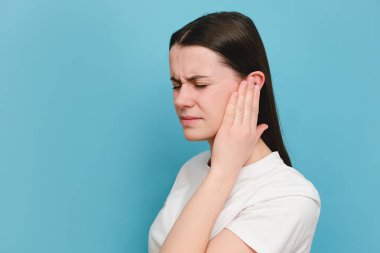 Close up of upset sad young millennial female holding painful ear, suddenly feeling strong ache, posing isolated on blue color background studio with copy space for advisement. Health problem concept clipart