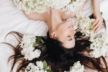 Güzel, sevimli, gülümseyen bir kadın portresi. Mükemmel cilt makyajı. Baharda ya da yaz çiçekleriyle rahat bir yatakta yatıyor. Güzel esmer kız doğal temizlik malzemesi. Saç Bakımı konsepti