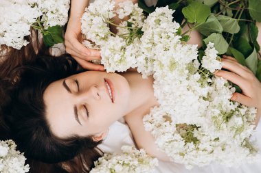 Romantik gülümseyen güzel bin yıllık beyaz kadın güzellik prosedüründen sonra temiz cilt ile yaz taze beyaz çiçekleriyle yatakta yatıyor. Cilt güzellik konsepti. Vücut parçası ve doğa
