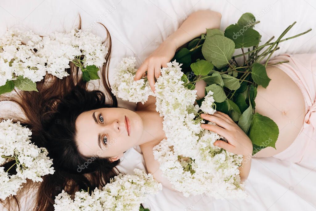 Portrait of calm young pregnant female with perfect skin, lying on comfortable white bed with beautiful fresh spring flowers, looking at camera. Concept motherhood. Future mother's expectation