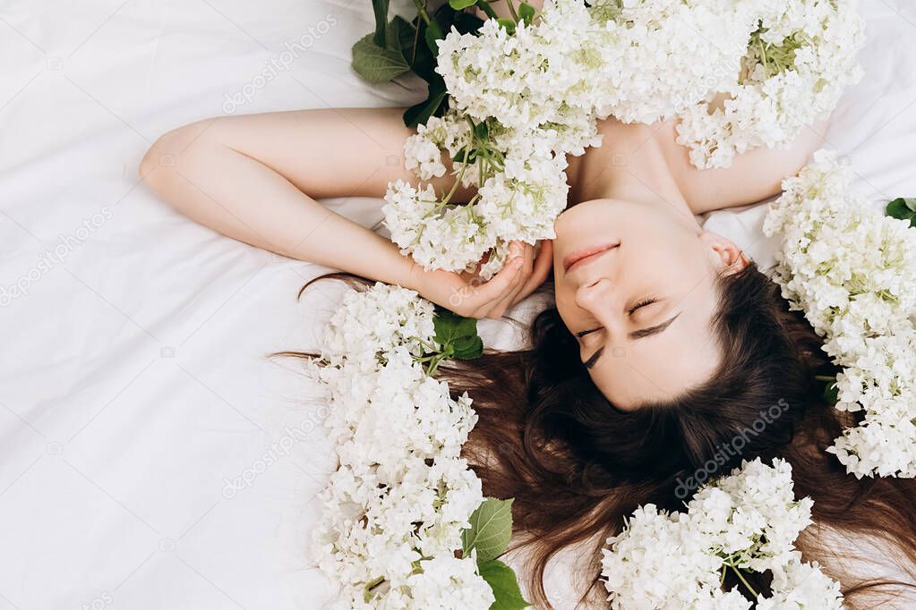 Portrait of dreamful woman with luxurious hair lying on cozy bed near white summer flowers around her head. Cheerful cute girl with eyes closed feel peaceful breathing fresh air, good pleasant smell