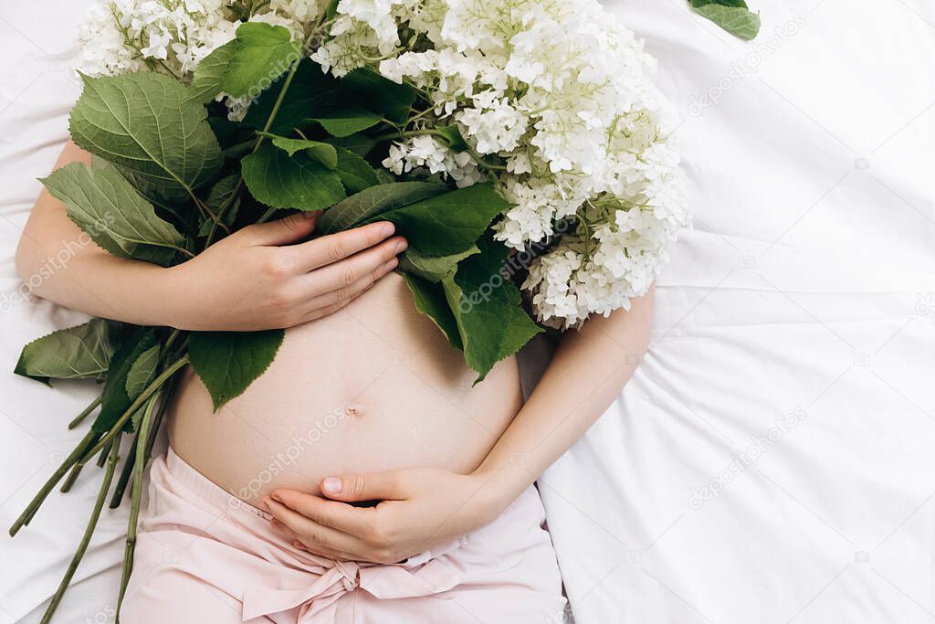 Close up of unrecognizable pregnant woman hands touching gently tummy lying on comfortable bed holding beautiful fresh summer flowers. Future mother's expectation. Copy space. Concept motherhood