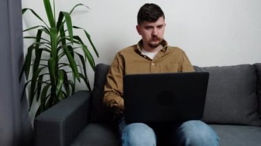 Ciddi genç öğrenci, evdeki gri koltukta dizüstü bilgisayarla oturuyor. Çevrimiçi iş konsepti. Odaklanmış girişimci uzaktan çalışıyor, bilgisayarda yazıyor, uzaktan yazışmalara öncülük ediyor.