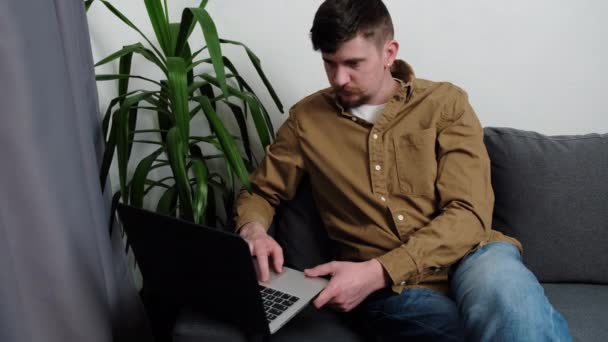 在笔记本电脑键盘上打字的年轻人坐在家里灰色的沙发上 看起来很沉思 专心致志地上网或在网上聊天 有胡子的家伙在无线电脑上学习 网上冲浪的概念 — 图库视频影像